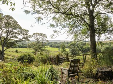 A garden area with wooden chairs under trees overlooking fields and trees at West Burnbank in Tarset near Bellingham