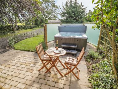 An outdoor patio with a wooden table and two chairs next to a hot tub with steps surrounded by greenery at Hen House View in Rugeley