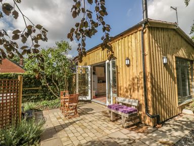 A wooden exterior building with open glass doors leading to a kitchen and a patio area with a wooden table and chairs and a bench with purple cushions at Hen House View in Rugeley