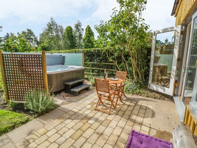 An outdoor patio with a hot tub a small wooden table and two chairs next to an open glass door at Hen House View in Rugeley