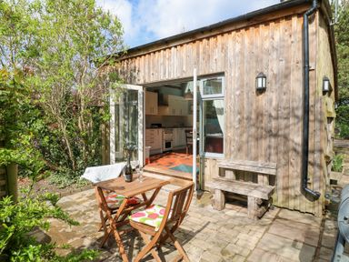 A wooden patio with a small table and two chairs with colorful cushions outside a wooden house with an open door showing a kitchen at Hen House View in Rugeley