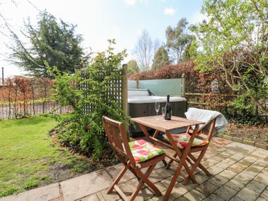 A small outdoor patio with a wooden table and two chairs with cushions next to a hot tub and garden at Hen House View in Rugeley