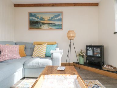 A living room with a light blue sofa multiple cushions a wooden coffee table a lamp a black stove and a painting on the wall at The Lily Pad in Camborne