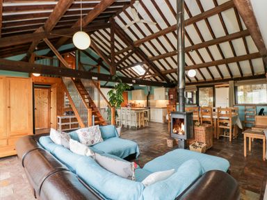 An open living area with a blue sectional sofa a wood burning stove a wooden dining table and chairs and a kitchen with white cabinets at The Barn in Draycot Cerne near Malmesbury