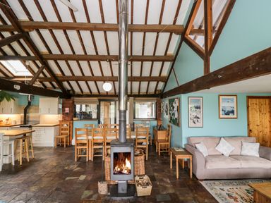 An open plan room with a wood stove in the center wooden dining chairs a kitchen area with stools and a sofa with cushions at The Barn in Draycot Cerne near Malmesbury