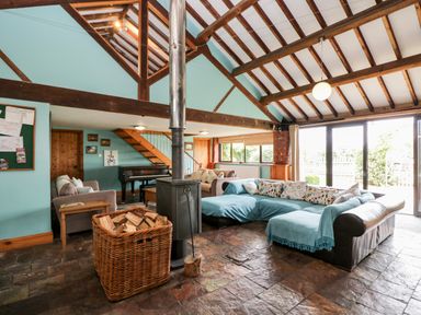 A living room with a wood stove and basket of firewood a sectional sofa piano and wooden stairs at The Barn in Draycot Cerne near Malmesbury