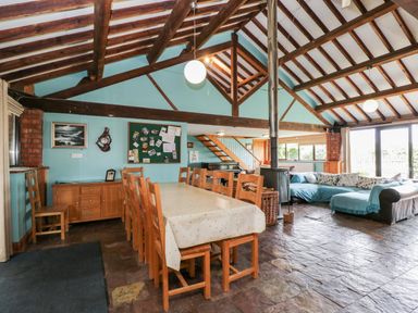 A dining area with a long table and wooden chairs next to a living area with a blue sofa and a wood stove under a beamed ceiling at The Barn in Draycot Cerne near Malmesbury