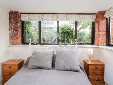 A bedroom with a bed between two wooden nightstands with lamps and windows behind at The Barn in Draycot Cerne near Malmesbury