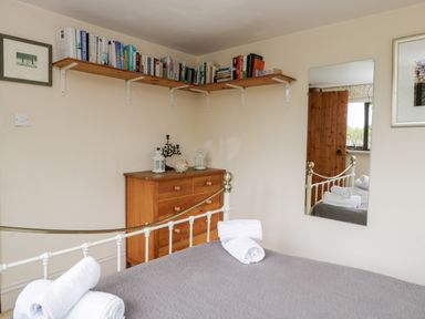 A bedroom with a metal bed frame with gray bedding and folded white towels on the bed a wooden chest of drawers and a wall shelf with books and decorations a mirror and two framed pictures on the walls at The Barn in Draycot Cerne near Malmesbury