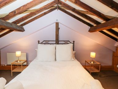 A bedroom with a double bed with white bedding and pillows under a ceiling with exposed wooden beams and two bedside tables with lamps at The Barn in Draycot Cerne near Malmesbury