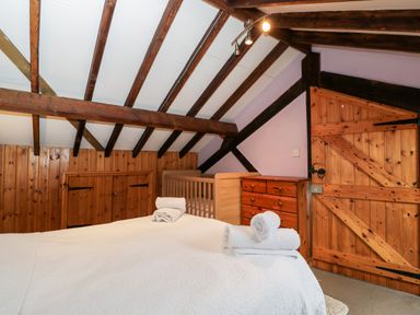 A bedroom with a bed covered in a white quilt and rolled towels wooden beams a crib a chest of drawers and a wooden door at The Barn in Draycot Cerne near Malmesbury