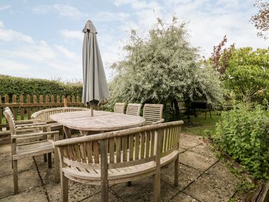 An outdoor patio with wooden table and chairs with a closed umbrella surrounded by trees and plants at The Barn in Draycot Cerne near Malmesbury
