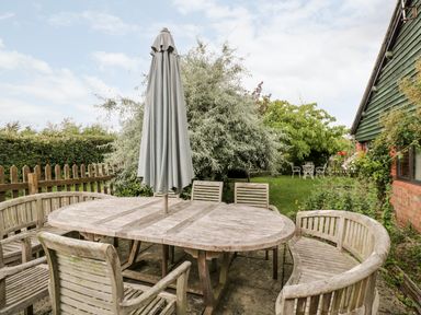 An outdoor wooden dining table with chairs and a closed umbrella in a garden next to a fenced lawn at The Barn in Draycot Cerne near Malmesbury