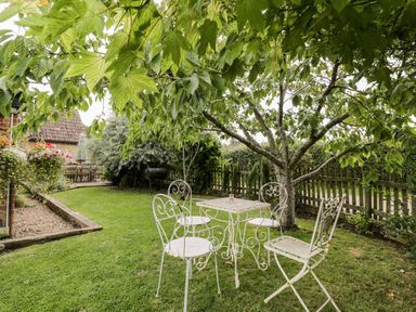 A garden with a white metal table and four chairs under a tree near a wooden fence at The Barn in Draycot Cerne near Malmesbury