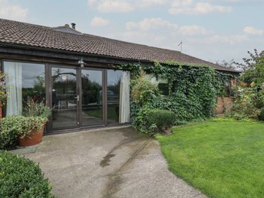 The exterior of a single-story building with large glass doors surrounded by greenery and plants at The Barn in Draycot Cerne near Malmesbury