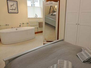 A bedroom with a gray bedspread and towels on the bed a bathtub near a window and a standing mirror a white wardrobe and a wall picture at Seion Chapel in Beaumaris