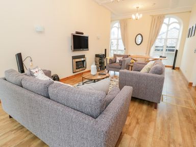 A living room with grey sofas a round wooden coffee table a wall-mounted television and large arched windows at Seion Chapel in Beaumaris