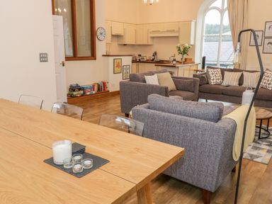 An open living and dining area with grey sofas a wooden dining table kitchen cabinetry and large arched window at Seion Chapel in Beaumaris