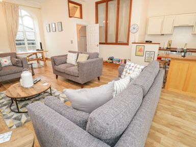 A living room with gray sofas a wooden coffee table a rug a wooden desk large window and a kitchen area at Seion Chapel in Beaumaris