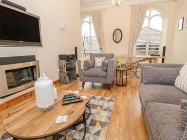 A living room with gray armchairs a wooden coffee table a wall-mounted television and large arched windows at Seion Chapel in Beaumaris