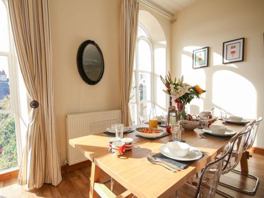 A dining room with a wooden table set for a meal transparent chairs and large windows with striped curtains at Seion Chapel in Beaumaris