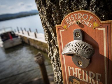A metal bottle opener on a wooden plaque mounted on a tree near a dock and water