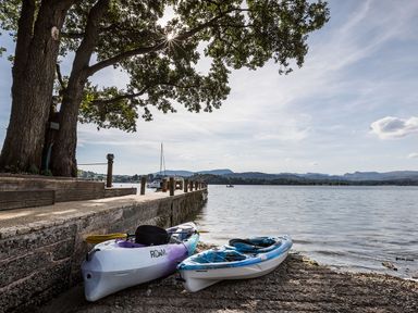 Two kayaks on a pebbled shore next to a wooden dock and large tree by a lake with mountains in the background at Waterhead Cottage Ecclerigg near Troutbeck Bridge