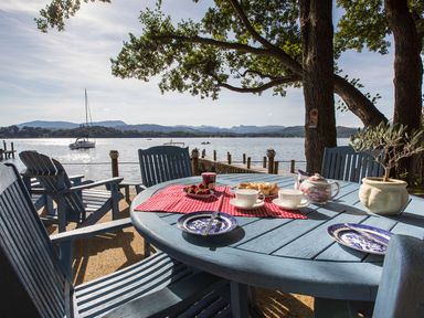 A lakeside patio with blue wooden chairs and table set for tea near a dock and sailboats