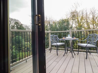 An outdoor balcony with a small metal table and two chairs with cushions near trees and bushes at Hazel Cottage in Poughill near Bude