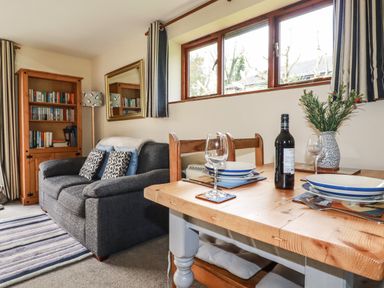 A living room with a dining table set with plates glasses a bottle and a sofa with cushions and a bookcase at Hazel Cottage in Poughill near Bude