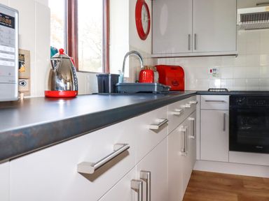 A kitchen with white cabinets a black countertop a red kettle a red bread box and a built in oven at Hazel Cottage in Poughill near Bude