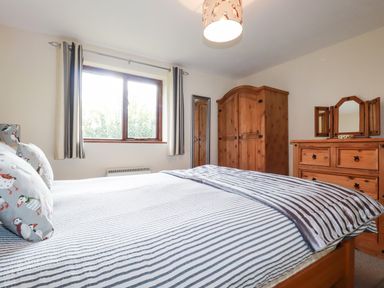 A bedroom with a bed striped bedding a wooden wardrobe a wooden dresser with a tri-fold mirror and a window with curtains at Hazel Cottage in Poughill near Bude