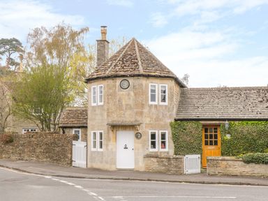 A stone house with a conical roof a white door and clock on the wall surrounded by a stone wall and greenery at Pike Cottage in Acton Turville near Chipping Sodbury