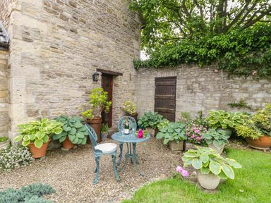 A small outdoor seating area with a round metal table and two chairs surrounded by potted plants next to a stone wall at Pike Cottage in Acton Turville near Chipping Sodbury