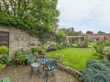A garden with a metal table and chairs on gravel next to a lawn and a stone wall with plants at Pike Cottage in Acton Turville near Chipping Sodbury