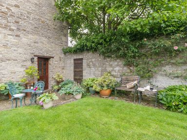 A garden with green grass a stone wall potted plants a bench and a metal table with chairs at Pike Cottage in Acton Turville near Chipping Sodbury