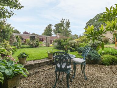 A garden with two metal chairs and a small table on gravel near various plants and a grassy lawn at Pike Cottage in Acton Turville near Chipping Sodbury