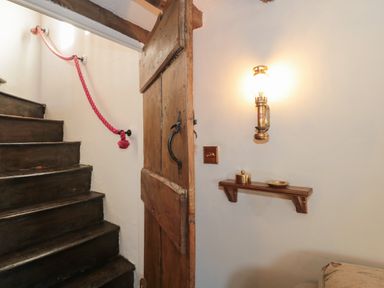 A wooden staircase with a red rope handrail beside a wooden door with a metal handle and a wall mounted light above a small wooden shelf at Pike Cottage in Acton Turville near Chipping Sodbury