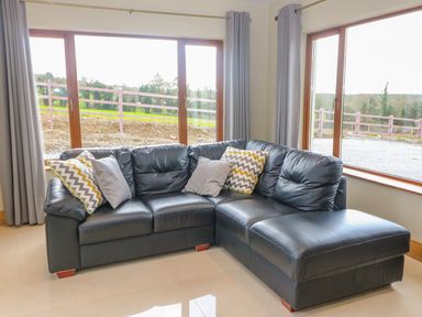 A corner black leather sofa with cushions in a room with large windows and grey curtains at Rossanean in Farranfore near Killarney County Kerry