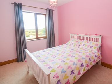 A bedroom with a white bed with colorful heart patterned bedding a window with gray curtains and pink walls at Rossanean in Farranfore near Killarney County Kerry
