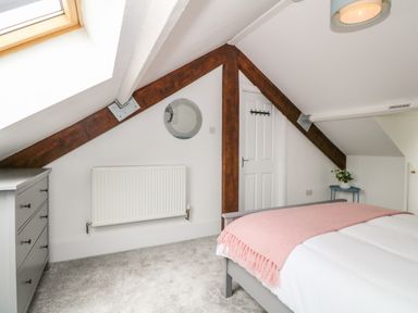 A bedroom with a bed covered by a pink throw, a grey chest of drawers, a small round table with a plant, exposed wooden beams, and a skylight window at Lyngaran House in Holyhead