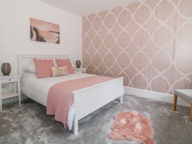 A bedroom with a white bed and pink bedding two bedside tables with lamps a chair a wall painting and patterned wallpaper at Lyngaran House in Holyhead