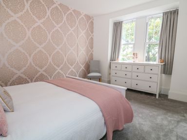 A bedroom with a bed a pink blanket a white dresser a chair and two windows with gray curtains at Lyngaran House in Holyhead