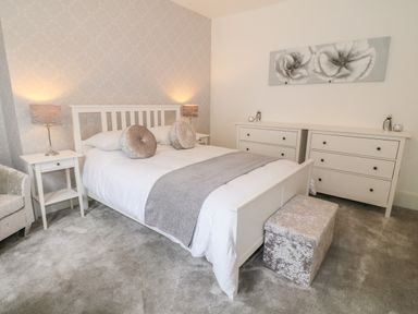 A bedroom with a white bed and grey cushions a white dresser with drawers and a grey ottoman at Lyngaran House in Holyhead
