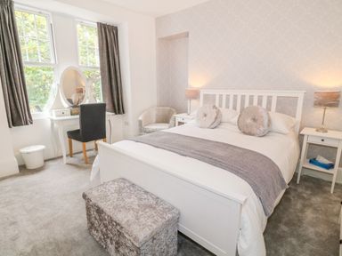 A bedroom with a white bed two round pillows gray throw two bedside tables with lamps a gray chair white dressing table with mirror and black chair at Lyngaran House in Holyhead