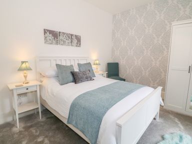 A bedroom with a white bed, bedside tables with lamps, a chair, a wardrobe, and patterned wallpaper at Lyngaran House in Holyhead