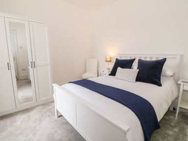 A bedroom with a white bed frame blue pillows white bedding a white wardrobe with a mirror and a white chair at Lyngaran House in Holyhead