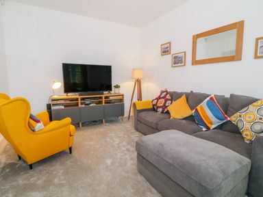 A living room with a grey sofa and yellow armchair facing a TV on a wooden stand at Lyngaran House in Holyhead