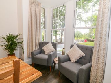 A sitting area with two gray armchairs with cushions next to large windows and a wooden table with a plant at Lyngaran House in Holyhead