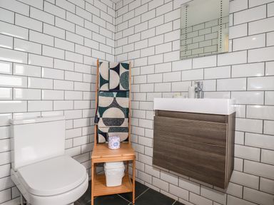 A bathroom with a toilet a wooden towel rack with patterned towels a small wooden shelf under the towel rack a floating sink with storage and a mirror on a tiled wall at Blue Bay Beach House in Mawgan Porth